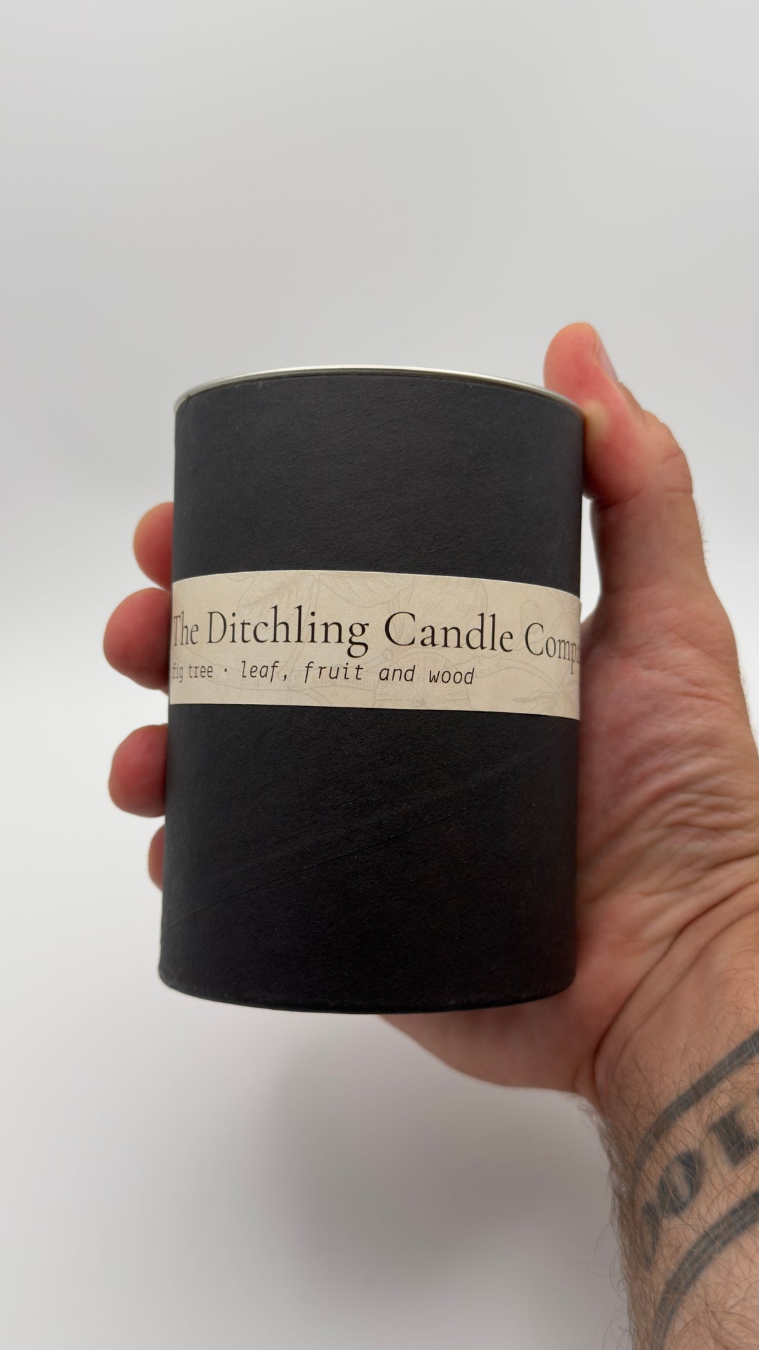 Hand holding a black cylindrical box for The Ditchling Candle Company Fig Tree candle, scented with leaf, fruit and wood.