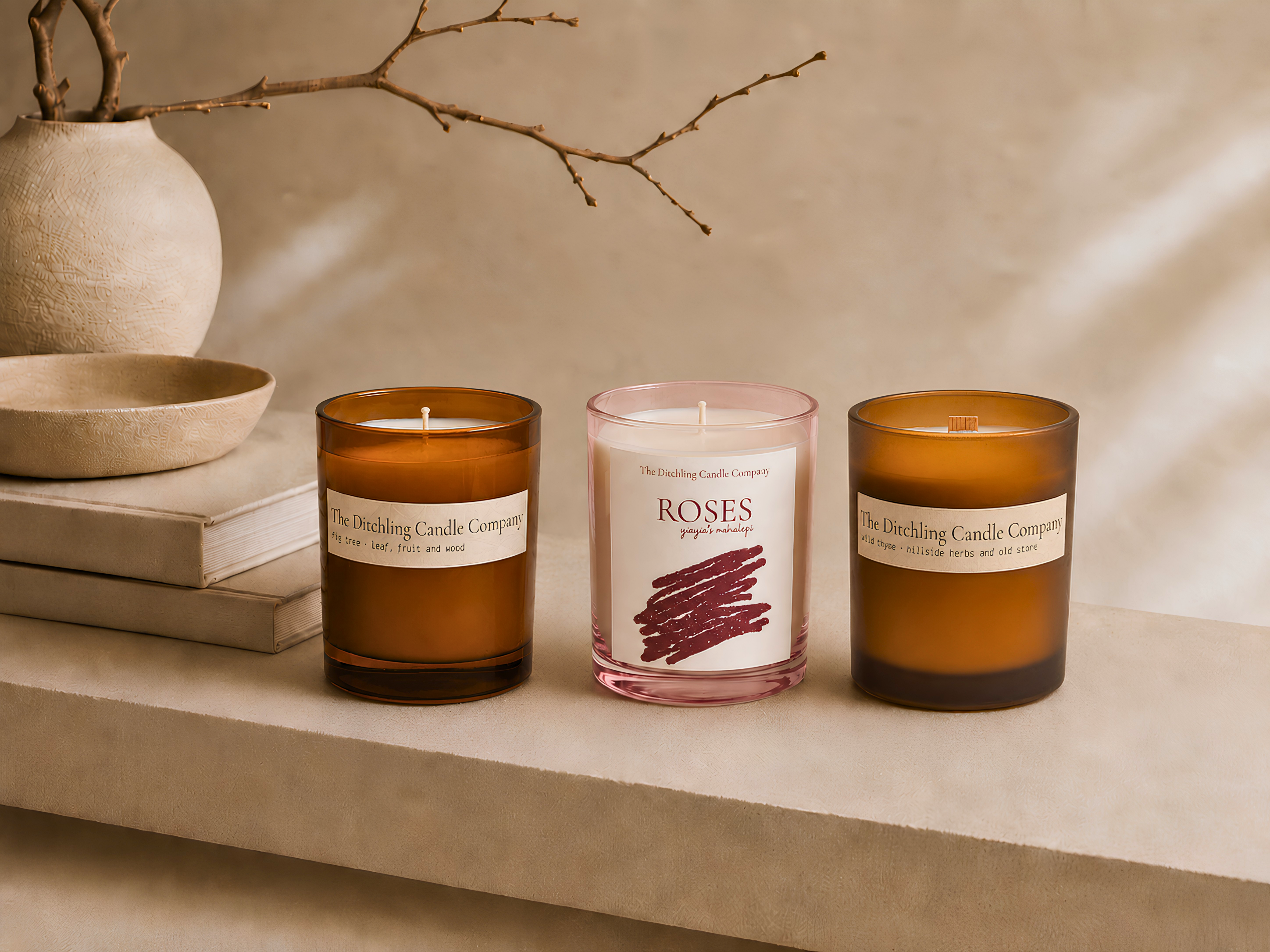 Three Ditchling Candle Company candles displayed on a neutral stone shelf: Fig Tree and Wild Thyme in amber glass, with Roses in pink glass at the centre.