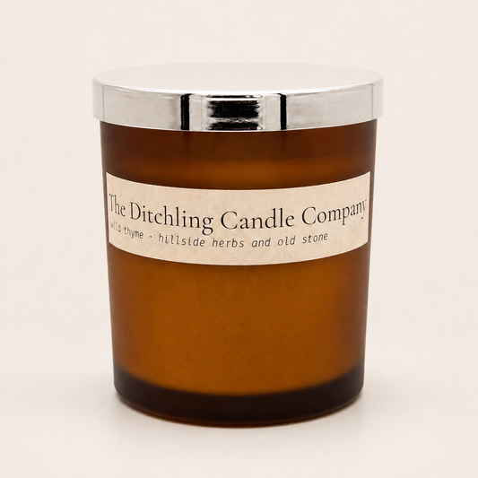 Amber glass wild thyme scented soy candle by The Ditchling Candle Company with a silver lid, shown on a warm off-white background.