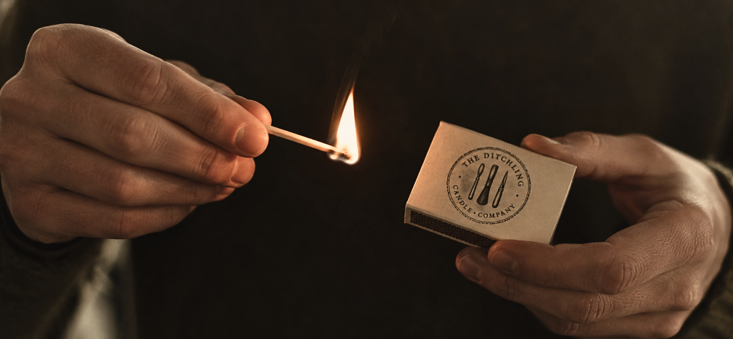 A person holds a lit match in one hand and a small matchbox in the other. The matchbox is cream-colored with a circular black logo reading 'THE DITHCLING CANDLE COMPANY'